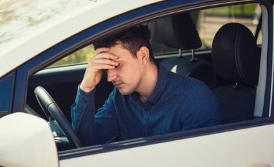 Casual young guy driver experiencing headache, should stop the car after driving in a traffic jam at rush hour. Exhausted, overloaded man keeps hand to head, healthcare concept, feeling anxiety.